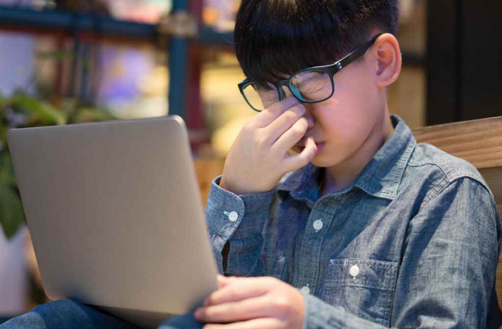 A boy squeezes his nose as his eyes is strained, stressed from long screen exposure from blue light  from his laptop computer,